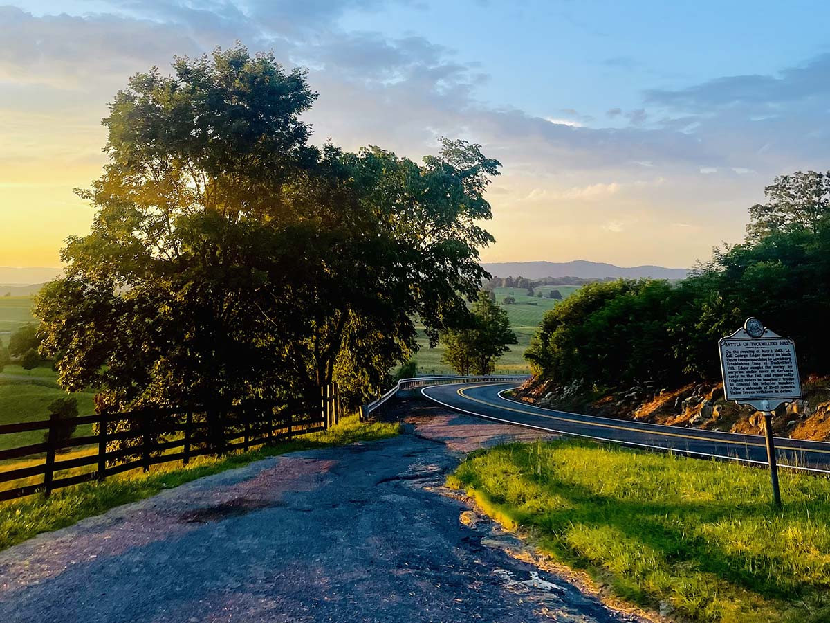 Scenic Route 60 leads through lush greenery under a vibrant sky at sunset, with a historical marker visible roadside.