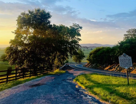 Scenic Route 60 leads through lush greenery under a vibrant sky at sunset, with a historical marker visible roadside.