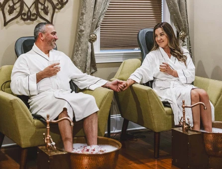 A couple enjoying a foot soak at Pomona Salt Cave and Spa--they are in white robes, seated in green chairs, enjoying drinks.