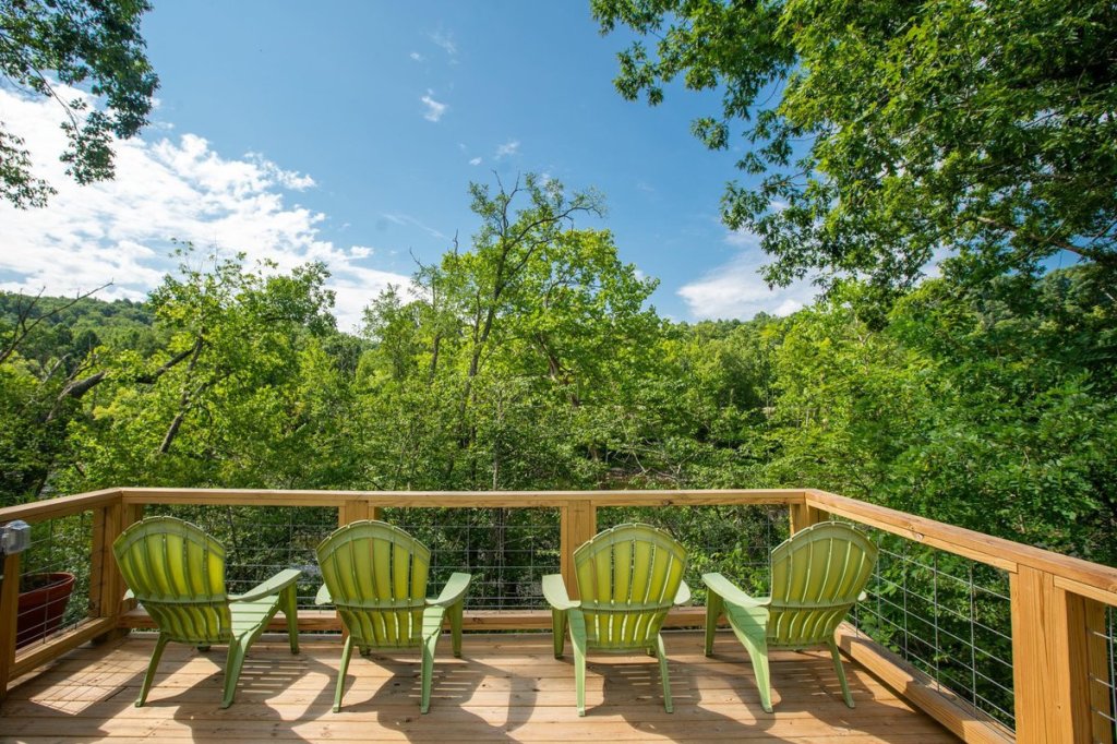 A wooden deck at the Riverstead features four green Adirondack chairs overlooking a lush forest with vibrant trees under a clear blue sky.